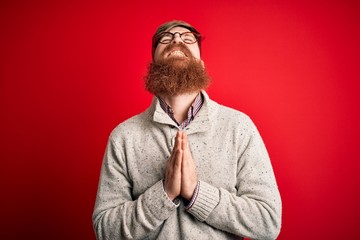 Handsome Irish redhead man with beard wearing casual sweater and glasses over red background...