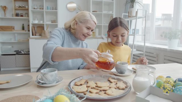Joyous grandmother pouring tea into cup for little granddaughter while sitting together at kitchen table with rabbit cookies and Easter eggs on them