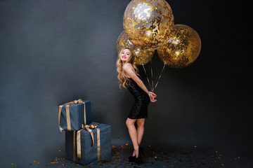 Beautiful stylish young woman on heels, long curly blonde, black luxury dress with big balloons full with golden tinsels on black background. Presents, birthday party, celebrating, smiling
