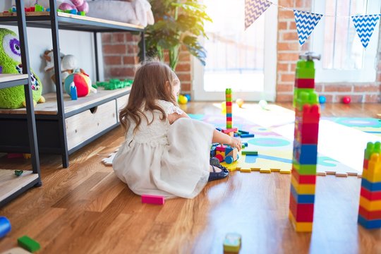 Adorable blonde toddler playing with building blocks around lots of toys at kindergarten