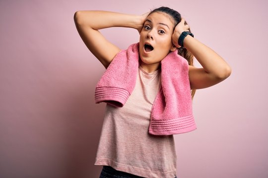 Young beautiful brunette sportswoman wearing sportswear and towel over pink background Crazy and scared with hands on head, afraid and surprised of shock with open mouth
