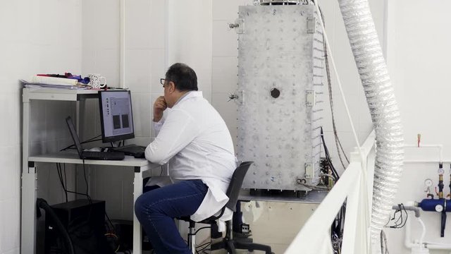 Rear view of a man engineer in white coat sitting at the table and working with a computer in metrological laboratory. Stock footage. Equipment and technologies concept.