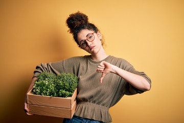 Young beautiful gardener woman with curly hair and piercing holding wooden box with plants with...