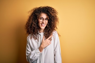 Young beautiful brunette woman with curly hair and piercing wearing shirt and glasses cheerful with a smile of face pointing with hand and finger up to the side with happy and natural expression