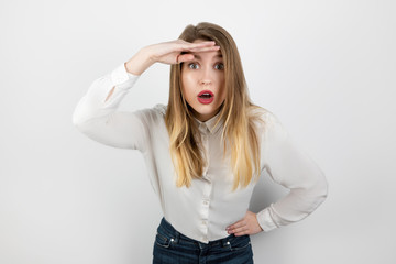 Fototapeta premium young curious blond woman holding her hand near her forehead gazing out standing on isolated white background