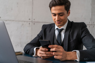 Smiling young businessman in dark suit using smartphone sitting in blurred office with dark blue table and laptop. Concept of good news and social media