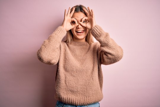 Young beautiful blonde woman wearing winter wool sweater over pink isolated background doing ok gesture like binoculars sticking tongue out, eyes looking through fingers. Crazy expression.