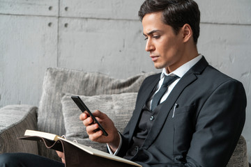 Side view of concentrated young businessman in dark suit using smartphone and holding notebook sitting in armchair in loft office. Corporate lifestyle concept