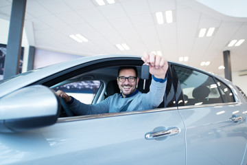 Middle aged smiling caucasian man sitting in brand new vehicle and showing keys to the camera at local vehicle dealership. Car purchase. Successful male buying new automobile.