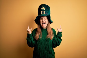 Fototapeta Young beautiful brunette woman wearing green hat on st patricks day celebration shouting with crazy expression doing rock symbol with hands up. Music star. Heavy concept. obraz