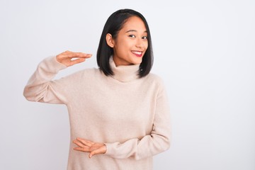 Young chinese woman wearing turtleneck sweater standing over isolated white background gesturing with hands showing big and large size sign, measure symbol. Smiling looking at the camera. Measuring