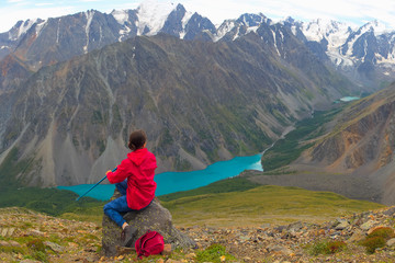 A girl in a red jacket is sitting on a high mountain. A woman is looking at a mountain lake. A tourist admires the panorama of the mountains.