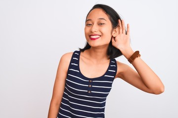 Young chinese woman wearing striped t-shirt standing over isolated white background smiling with hand over ear listening an hearing to rumor or gossip. Deafness concept.