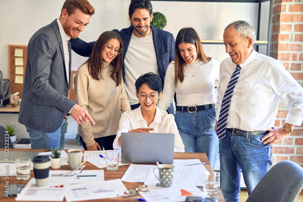 Wall mural Group of business workers smiling happy and confident. One of them sitting and partners standing around. Working together with smile on face looking at the laptop at the office