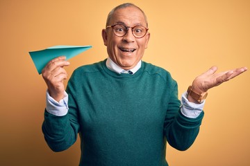 Senior handsome man holding airplane of paper standing over isolated yellow background very happy and excited, winner expression celebrating victory screaming with big smile and raised hands