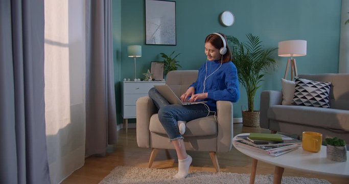 Caucasian young pretty woman in big white headphones sitting in the armchair at the window at home and listening to the music or watching funny video on laptop computer while typing on keyboard.