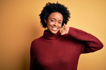 Fototapeta na wymiar Young beautiful African American afro woman with curly hair wearing casual turtleneck sweater smiling doing phone gesture with hand and fingers like talking on the telephone. Communicating concepts.