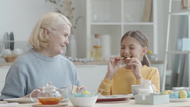 Playful little girl holding two bunny cookies in front of her eyes and then eating them while sitting together with cheerful grandma at kitchen table and having Easter dinner