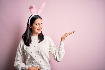 Young caucasian woman wearing cute easter rabbit ears over pink isolated background smiling cheerful presenting and pointing with palm of hand looking at the camera.