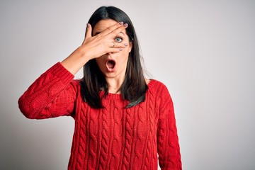 Young brunette woman with blue eyes wearing casual sweater over isolated white background peeking in shock covering face and eyes with hand, looking through fingers with embarrassed expression.