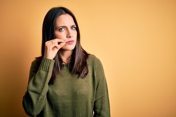 Young brunette woman with blue eyes wearing green casual sweater over yellow background mouth and lips shut as zip with fingers. Secret and silent, taboo talking