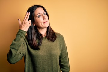 Young brunette woman with blue eyes wearing green casual sweater over yellow background Shooting and killing oneself pointing hand and fingers to head like gun, suicide gesture.