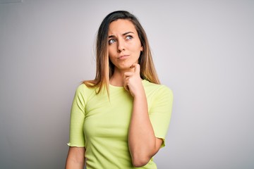 Beautiful blonde woman with blue eyes wearing green casual t-shirt over white background with hand on chin thinking about question, pensive expression. Smiling with thoughtful face. Doubt concept.