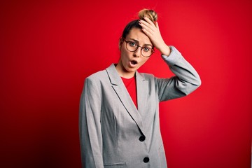 Young beautiful blonde businesswoman with blue eyes wearing glasses and jacket surprised with hand...