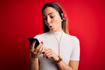 Young beautiful brunette woman listening to music using smartphone and headphones scared in shock with a surprise face, afraid and excited with fear expression