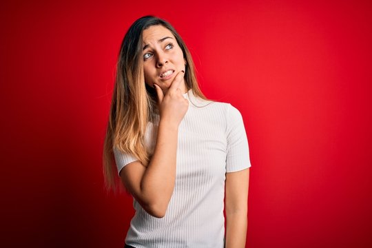 Beautiful blonde woman with blue eyes wearing casual white t-shirt over red background Thinking worried about a question, concerned and nervous with hand on chin