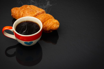 Heart pattern coffee cup In the cup there is black coffee and smoke Beside the coffee cup is the croissant.