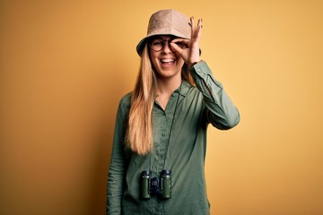 Beautiful blonde explorer woman with blue eyes wearing hat and glasses using binoculars doing ok gesture with hand smiling, eye looking through fingers with happy face.