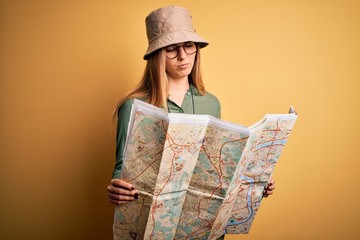 Blonde explorer woman with blue eyes on vacation wearing hat and binoculars holding city map with a confident expression on smart face thinking serious