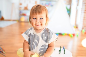 Adorable toddler standing with smile on face holding feeding bottle around lots of toys at kindergarten