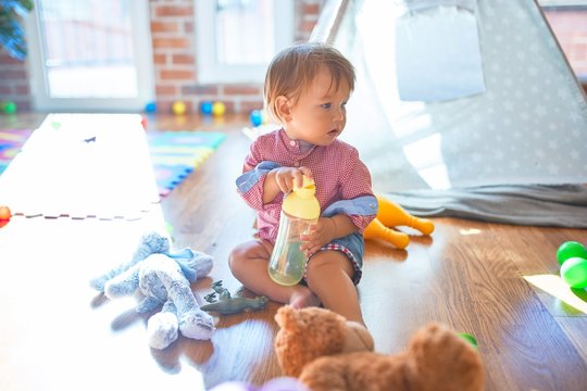 Adorable toddler holding feeding bottle around lots of toys at kindergarten