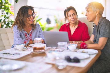 Meeting of middle age women having lunch and drinking coffee. Mature friends smiling happy using laptop at home on a sunny day