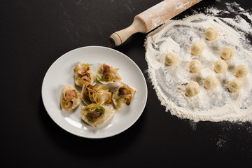 delicious varenyky with cracklings on plate near raw dough balls, flour and rolling pin on black background