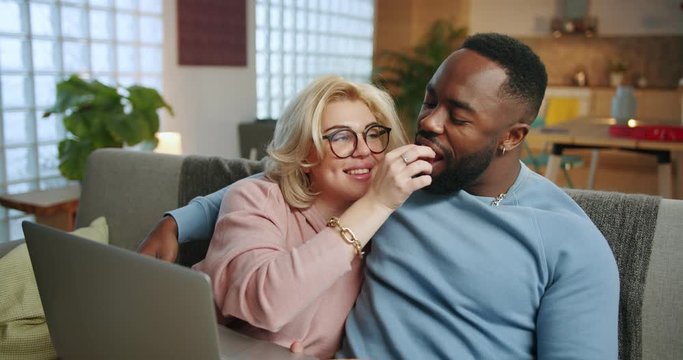 Entertainment time of pleasant mixed race couple resting on couch using laptop at home, romantic of family embracing and kissing each other, cute blonde woman feeding her boyfriend at cozy living room