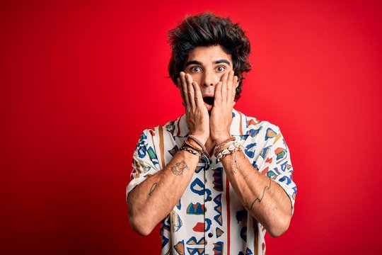 Young handsome man on vacation wearing summer shirt over isolated red background afraid and shocked, surprise and amazed expression with hands on face