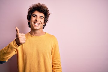 Young handsome man wearing yellow casual t-shirt standing over isolated pink background doing happy thumbs up gesture with hand. Approving expression looking at the camera showing success.