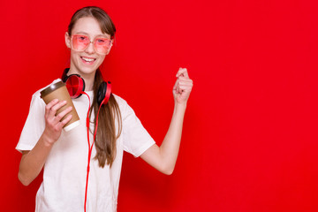 Amazed caucasian brunette teenage girl,holds coffe and wearing red earphones over red isolated background is happy and excited doing winner gesture with arms raised.Celebration concept.