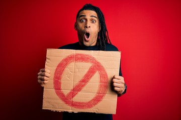 Young handsome african american man with dreadlocks holding banner with prohibited signal scared in...