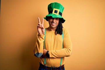 Young african american man wearing green hat celebrating saint patricks day smiling with happy face winking at the camera doing victory sign. Number two.