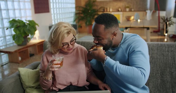 Engaging Afro-American boyfriend sharing tasty slice of pizza with beautiful blonde girl, joyful Caucasian girlfriend holding glass of beer and computer, mixed race couple enjoying moment at home
