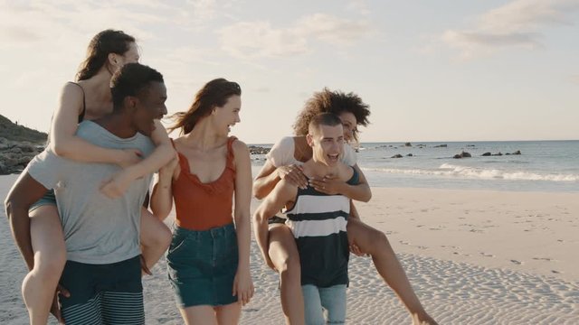 Two happy young men giving their girlfriends piggyback rides at the beach. Group of friends enjoying beach holidays.