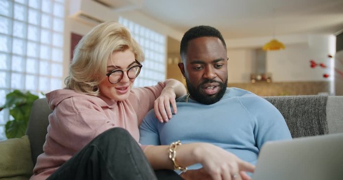 Happy mixed ethnicity young couple using laptop for e-commerce doing online shopping, together watching video movie sitting on sofa, smiling African man and Caucasian woman looking at computer at home