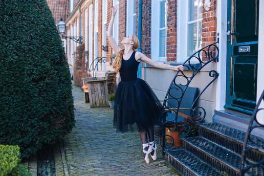 Young Ballerina Woman With Red Hair In Ballet Costume And Pointe Shoes Is In Beautiful Pose Dancing On Street. Ballerina Is Posing In The Streets. Girl Dancing Outdoors. Street Ballet And Performance