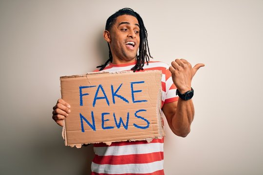 Young african american man with dreadlocks holding banner with fake news message protest pointing and showing with thumb up to the side with happy face smiling