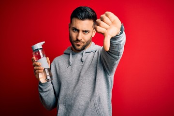 Young handsome sportsman drinking bottle of water to refeshment over red background with angry...