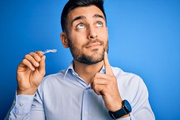 Young handsome man holding dental aligner tooth correction over blue background serious face thinking about question, very confused idea